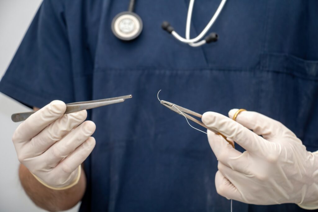 Doctor holds, in hand with disposable glove, forceps and needle holder and suture thread.